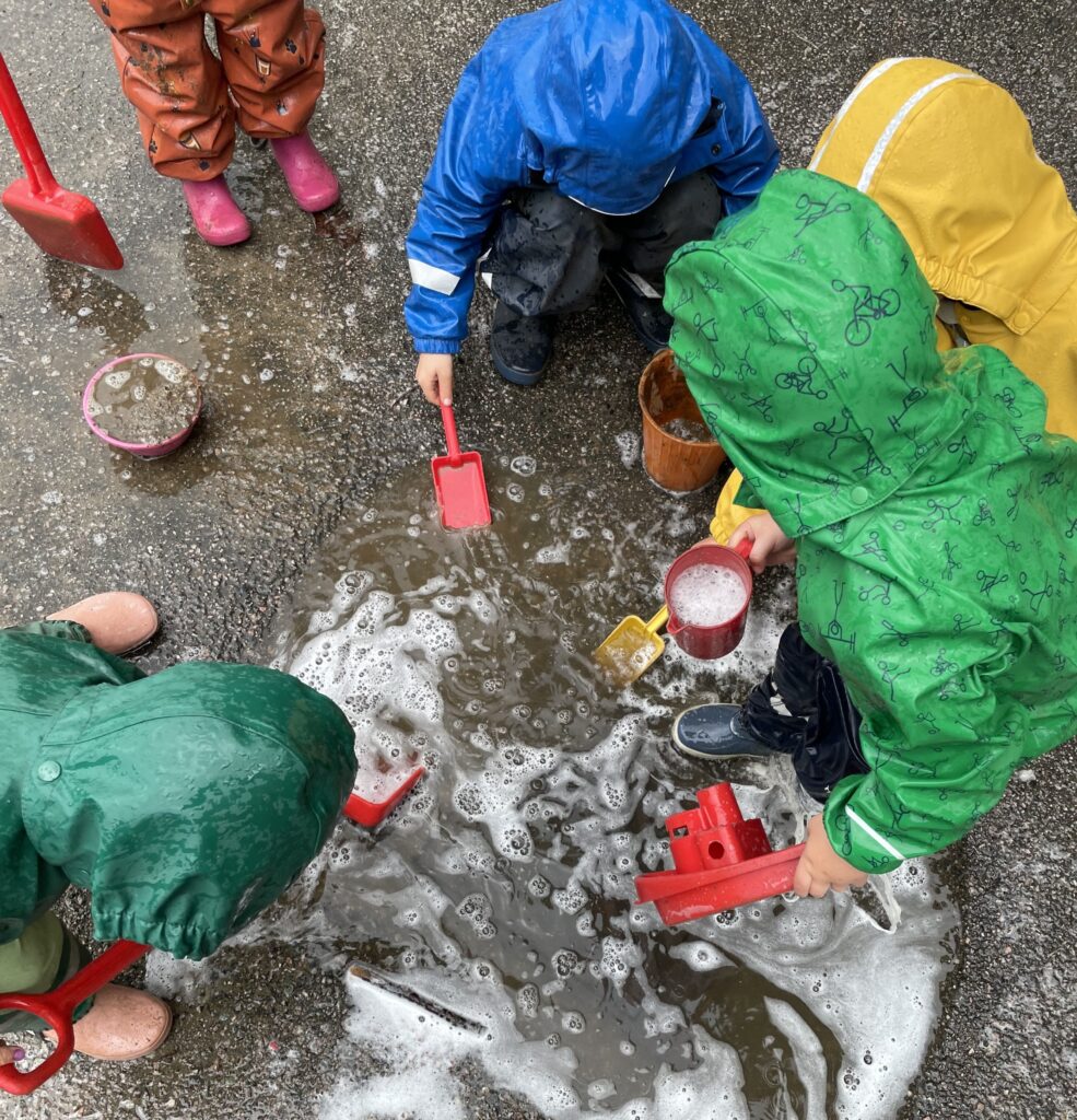 A small group of children dressed in raincoats and boots play with rainwater using plastic shovels, cups, boats and pail.